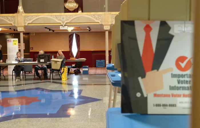 A voting center with poll workers seated at a table in the background and a sign in the foreground displaying voter information with a checkmark and a suit illustration, captured during Montana Election season. Blue voting booths line the right side of the image.